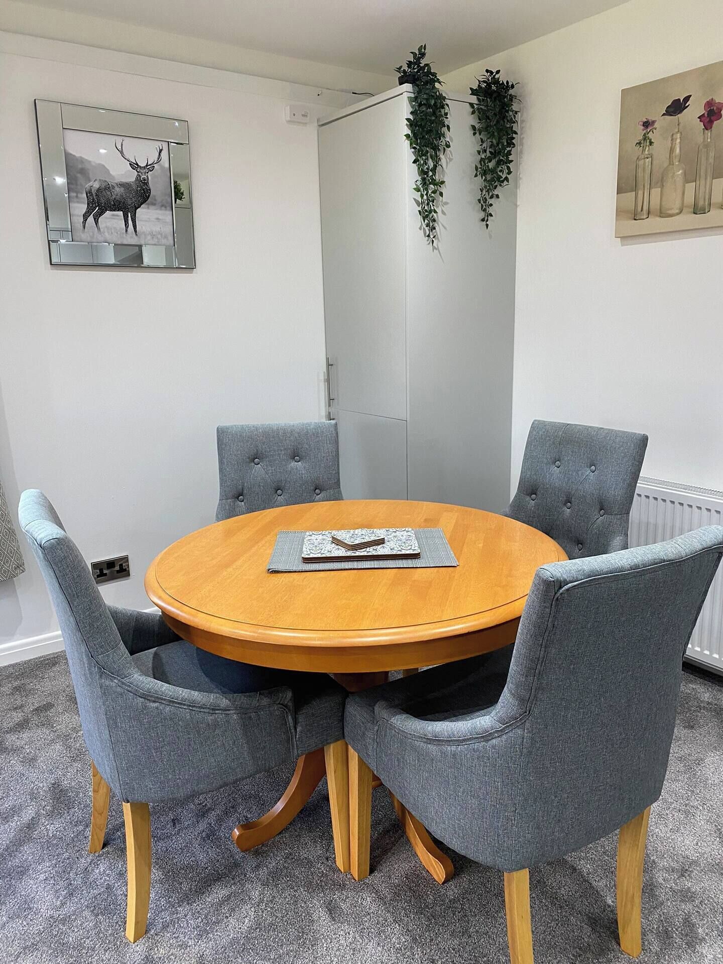 Une table ronde en bois, entourée de quatre chaises grises à revêtement rembourré, une œuvre d’art représentant un cerf, encadrée, accrochée au mur, et une plante en pot suspendue au-dessus.