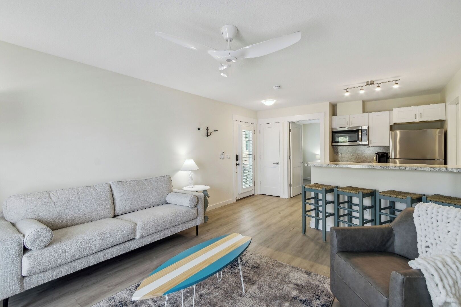A modern living room with a sofa, a coffee table, and a surfboard. There is a kitchen area with a microwave and a refrigerator.