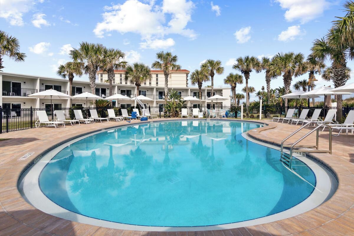 A pool area with lounge chairs and umbrellas, surrounded by palm trees and a building with multiple balconies.