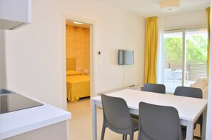 Una habitación de hotel compacta con kitchenette, una cama con ropa de cama amarilla, un televisor montado en la pared y un balcón con cortinas amarillas.