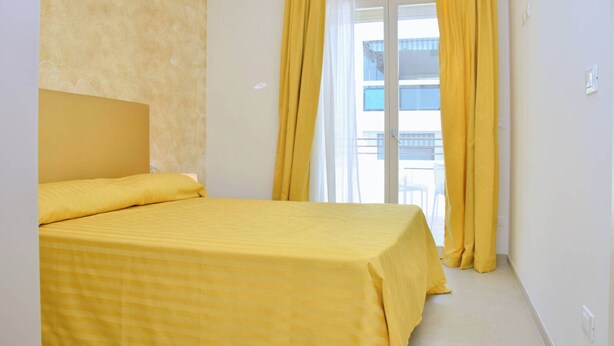A room with a bed, a window with yellow curtains, and a view of a building outside. - Sunny and Bright Beautiful Apartment (San Michele al Tagliamento)