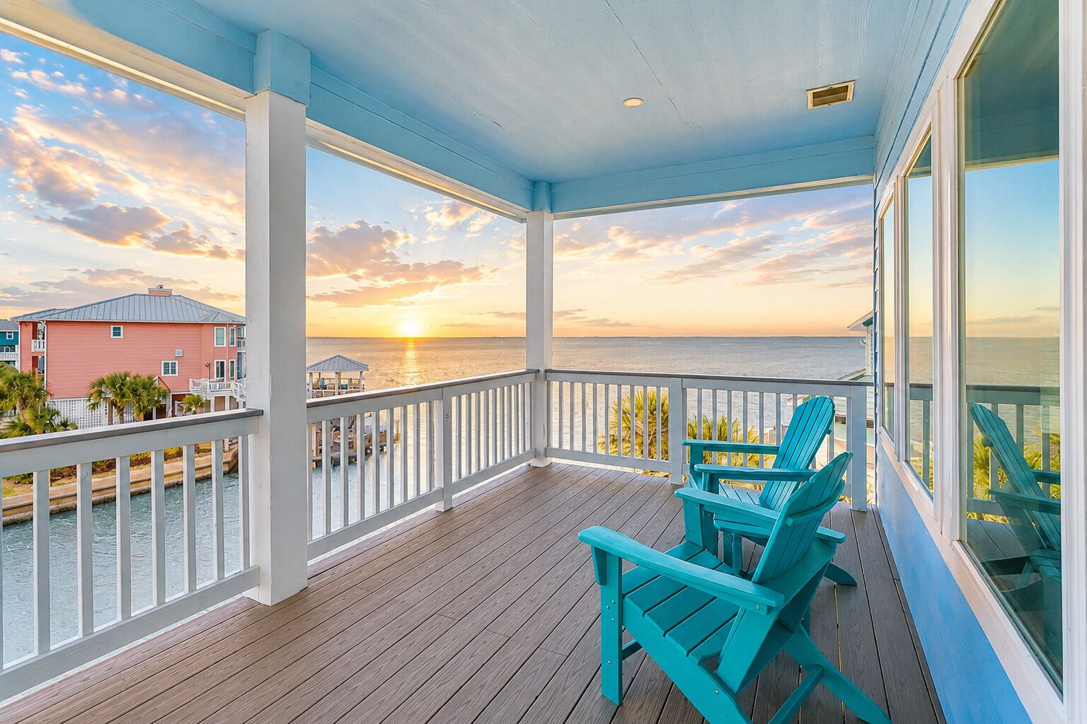 Un balcon abrité avec un plancher en bois, des rampes blanches et deux chaises turquoise.