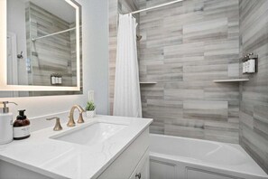 Un baño moderno con tocador blanco, un espejo grande, una bañera y una zona de ducha con cortina.