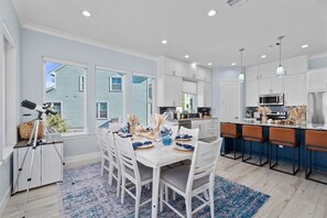 Una cocina moderna con armarios blancos, una isla central con taburetes y una zona de comedor con una mesa blanca dispuesta para cuatro personas.