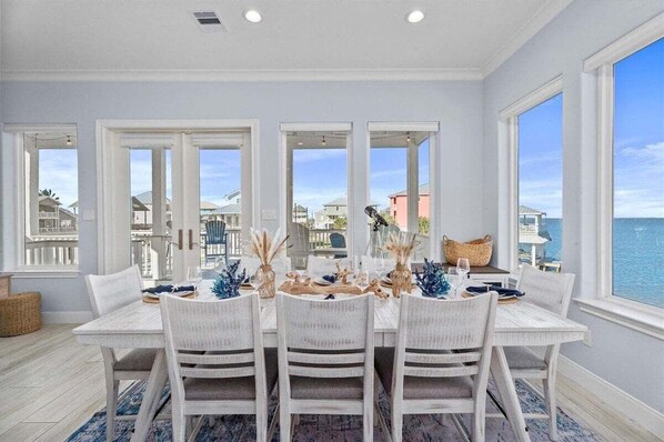 A dining area with a white table and chairs, large windows with a view of the sea and buildings, and a blue rug on the floor.