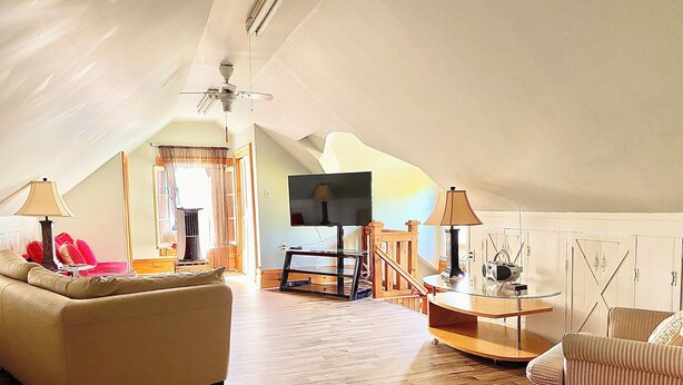 A spacious attic living room with a high ceiling, wooden flooring, a sofa, a chair, a TV, and a table with lamps.