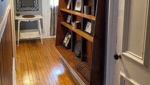 A hallway with wooden flooring, a white door, a white console table with a mirror, and a wooden bookcase with framed pictures and decorative items.
