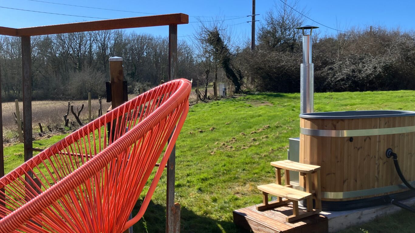 Uma banheira de hidromassagem de madeira com uma pequena mesa de madeira, uma cadeira vermelha e uma estrutura de pérgola em área externa gramada.