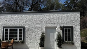 A small, single-story building with a white stucco exterior, two windows with shutters, a door, and a small porch area with potted plants.