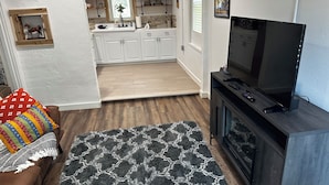 A living room with a brown sofa, a black and white rug, a TV on a wooden stand, and a kitchen in the background.