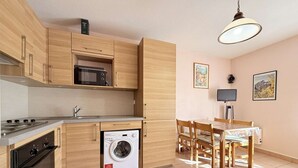 A compact kitchen with wooden cabinets, a microwave, a stove, and a washing machine. There is a dining table with chairs and a hanging light fixture. - C21Loctionvacances, T3 DUB- plages et commerces à pied (BORMES LES MIMOSAS)