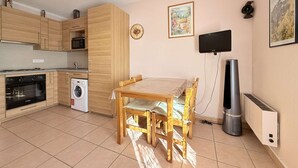 A compact kitchen with wooden cabinets, a microwave, oven, and washing machine. There is a dining table with chairs and a flat-screen TV mounted on the wall. - C21Loctionvacances, T3 DUB- plages et commerces à pied (BORMES LES MIMOSAS)