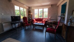 A living room with a red sofa, a coffee table, and a TV. There are windows with a view of trees outside. - T4 , Jardin arboré et plages à moins de 5 min à pieds (BORMES LES MIMOSAS)