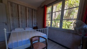 A bedroom with a bed, a chair, a window with curtains, and a standing fan. - T4 , Jardin arboré et plages à moins de 5 min à pieds (BORMES LES MIMOSAS)