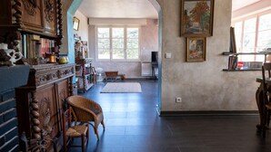 A spacious living room with a large wooden cabinet, a wicker chair, and a view through an archway to another room. - T4 , Jardin arboré et plages à moins de 5 min à pieds (BORMES LES MIMOSAS)