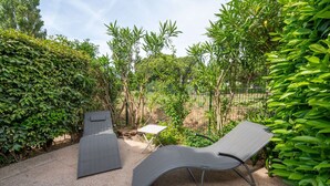 A patio with a curved lounge chair and a small table, surrounded by lush greenery and a fence.