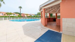 A swimming pool area with a covered seating area, palm trees, and a view of other buildings in the background.