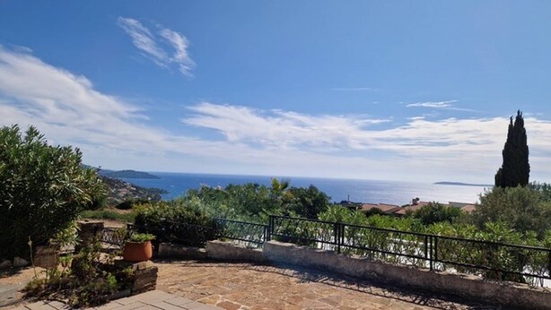 Une terrasse en pierre avec une rambarde en métal, offrant une vue sur un paysage côtier, un ciel dégagé et la mer à l’horizon.