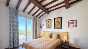 Une chambre avec un grand lit, des poutres apparentes, une vue à travers une porte coulissante et trois tableaux encadrés accrochés au mur.