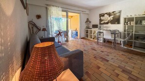 A living room with a sofa, a table, and a dining area with chairs. There is a window with curtains and a view of the outside. - T2 en rez de jardin , proche plage de St Clair et commerces (LE LAVANDOU)