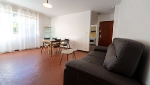 Un salon avec un canapé en cuir noir, une table à manger avec des chaises, un coin cuisine et une grande fenêtre ornée de rideaux légers.