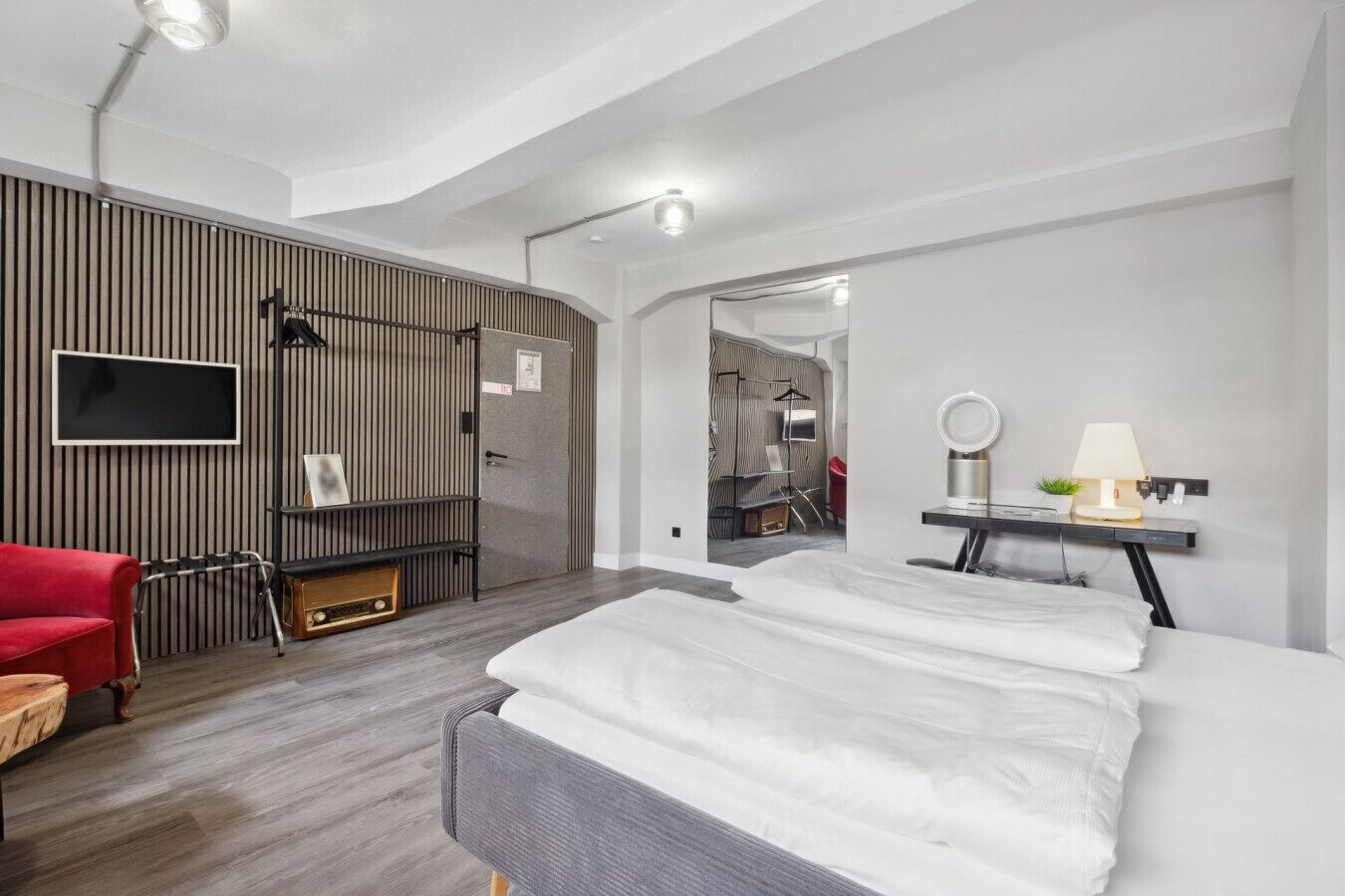 A modern hotel room with a bed, a red chair, a TV, a desk with a lamp, and a wardrobe.