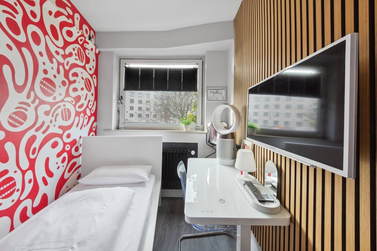 A hotel room with a bed, a desk with a mirror, a radiator, and a window with blinds.