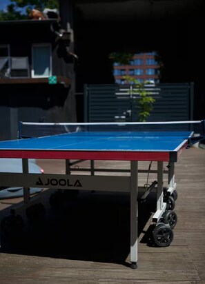 A table tennis table with a Joola brand logo, set up outdoors on a wooden deck with a building in the background. - The Room 44 (Hamburg)