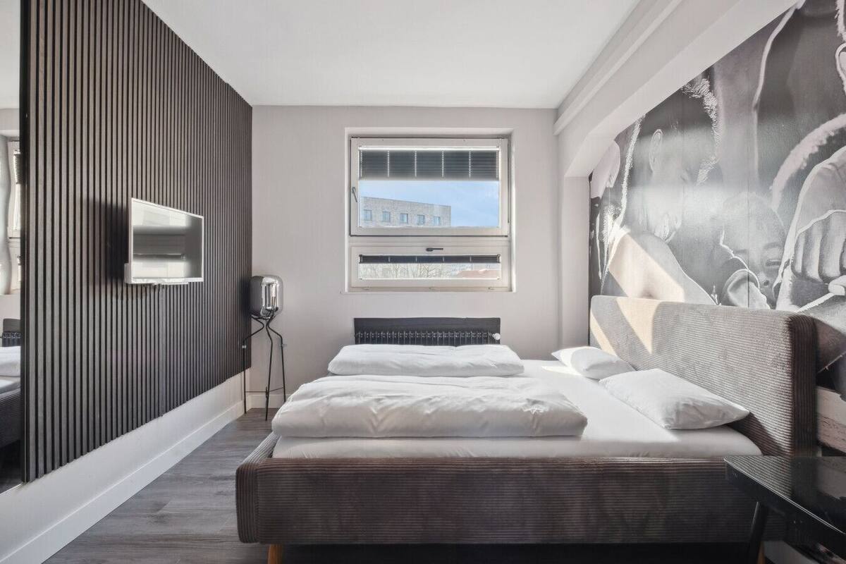 A modern hotel room with a bed, a window with blinds, a wall-mounted TV, and a large abstract painting.