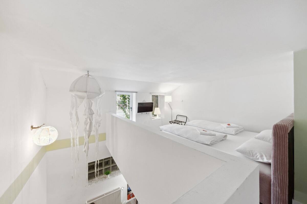 A white room with a bed, a hanging light fixture, a TV, and a window with a view of greenery.