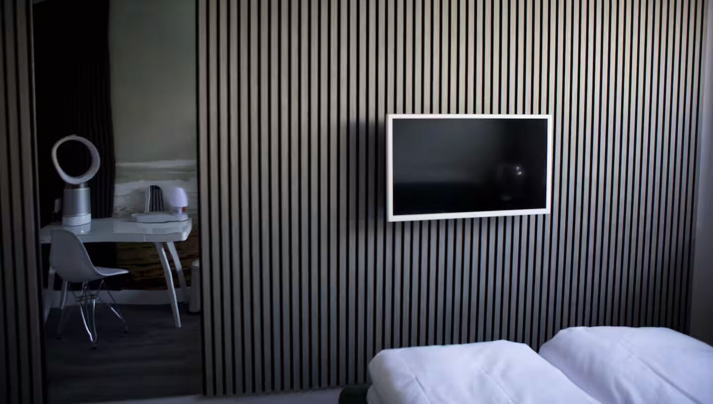 Habitación de hotel con una cama, un televisor montado en la pared, un escritorio con una silla y un tocador con espejo.