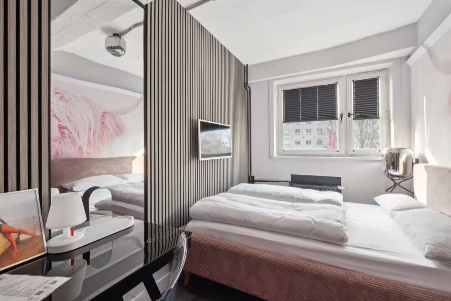 A modern hotel room with a large bed, a desk with a chair, a TV, and a window with blinds.