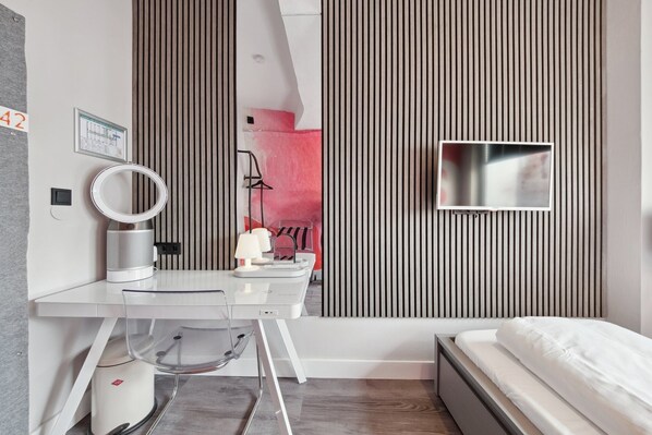 A modern hotel room with a white desk, a round mirror, a small TV, and a bed with white bedding. - The Room 44 (Hamburg)
