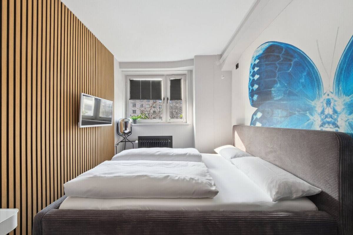A modern hotel room with a large bed, a television, a window with blinds, and a wall with vertical wooden slats.