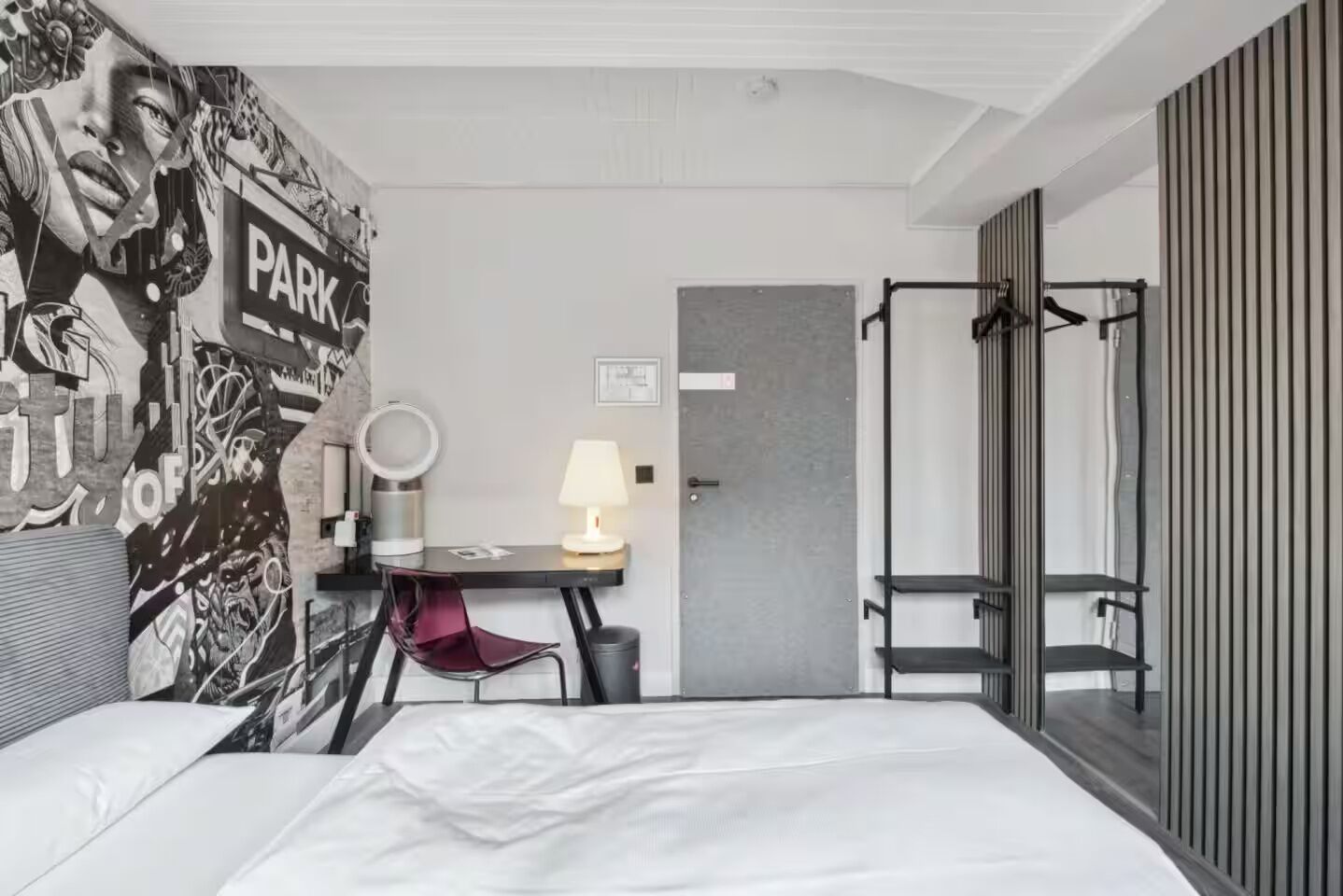 A hotel room with a bed, a desk with a lamp, a chair, a mirror, and a wardrobe.