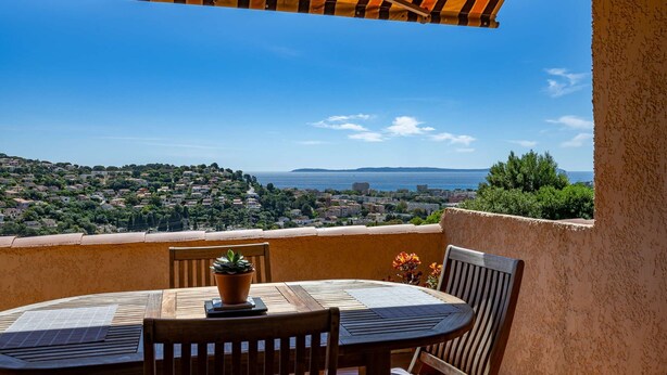 A terrace with a table and chairs, overlooking a coastal town and the sea.