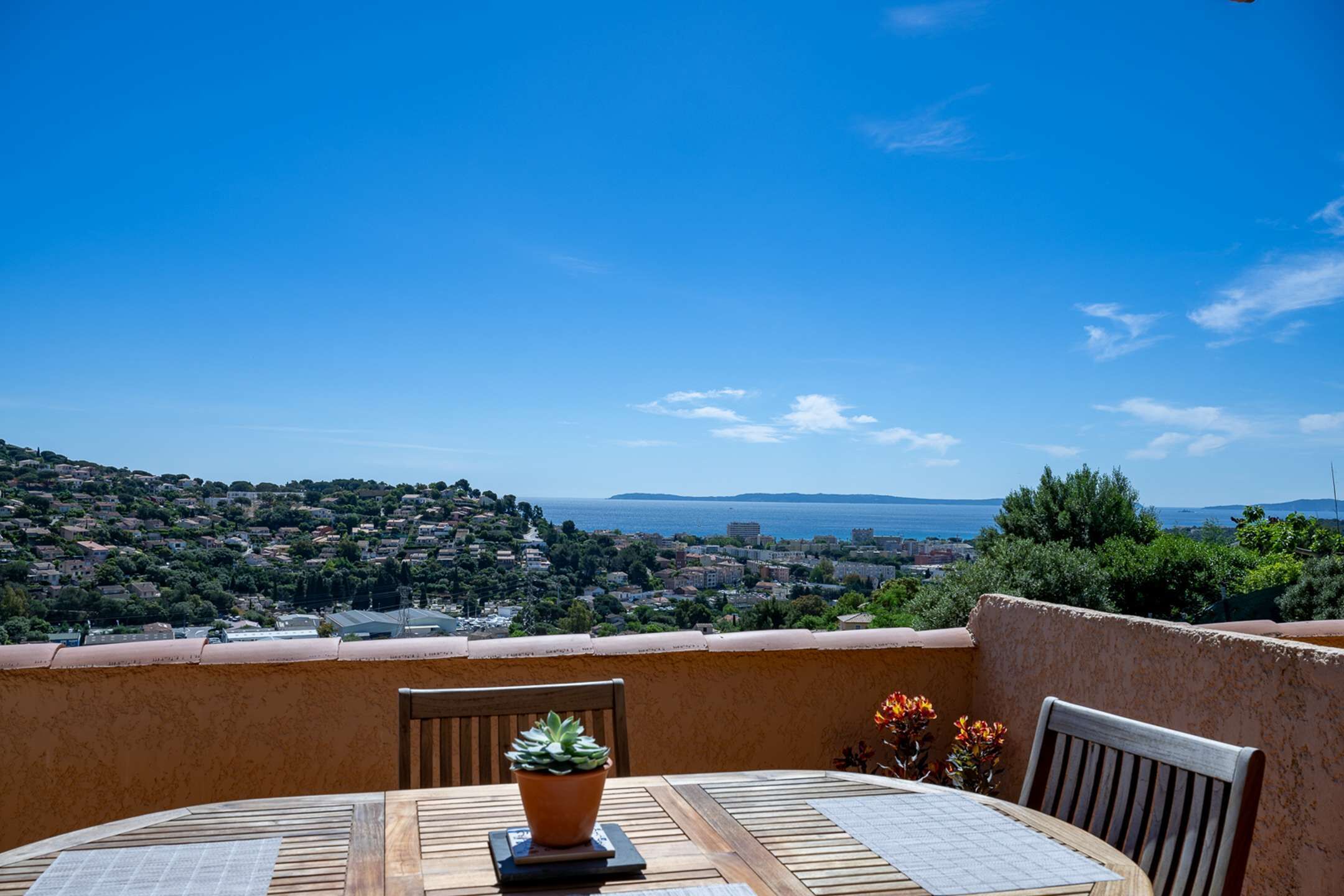 A round wooden table with a potted plant and two chairs, overlooking a coastal town with a clear blue sky.