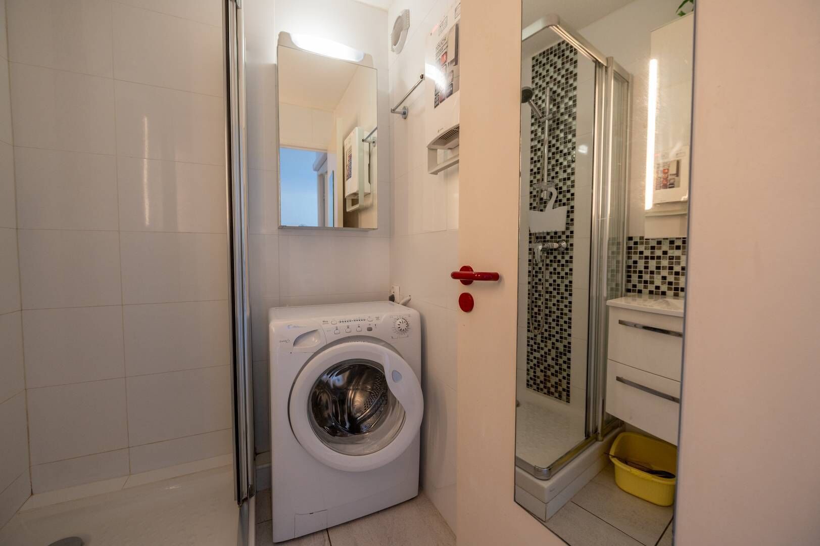 Ett litet badrum med tvättmaskin, en dusch med mosaik och en gul tvättkorg.