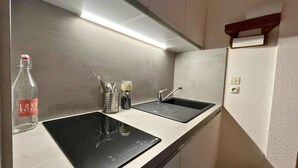 Une cuisine compacte dotée de deux plaques de cuisson à induction noires, d’un évier en acier inoxydable et d’un petit plan de travail. On y trouve une bouteille d’eau et un porte-ustensiles sur le plan de travail.
