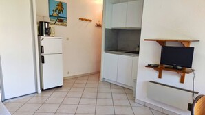 Une cuisine compacte équipée d’un réfrigérateur, d’un four à micro-ondes et d’un petit îlot central. Un téléviseur à écran plat est fixé sur une étagère en bois au-dessus de l’îlot.