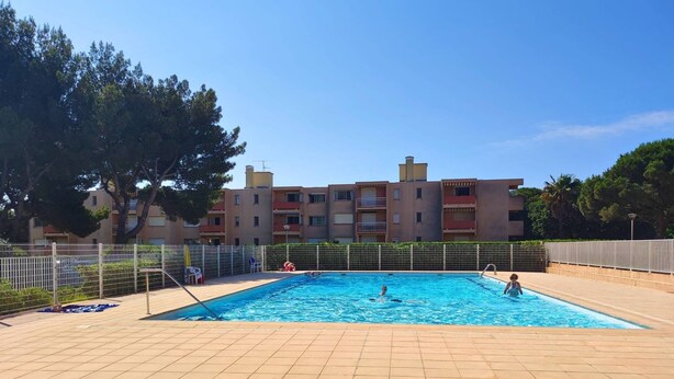 Une piscine dont le fond est recouvert de carrelage, entourée d’une clôture en métal, et un bâtiment avec de nombreux balcons en arrière-plan.