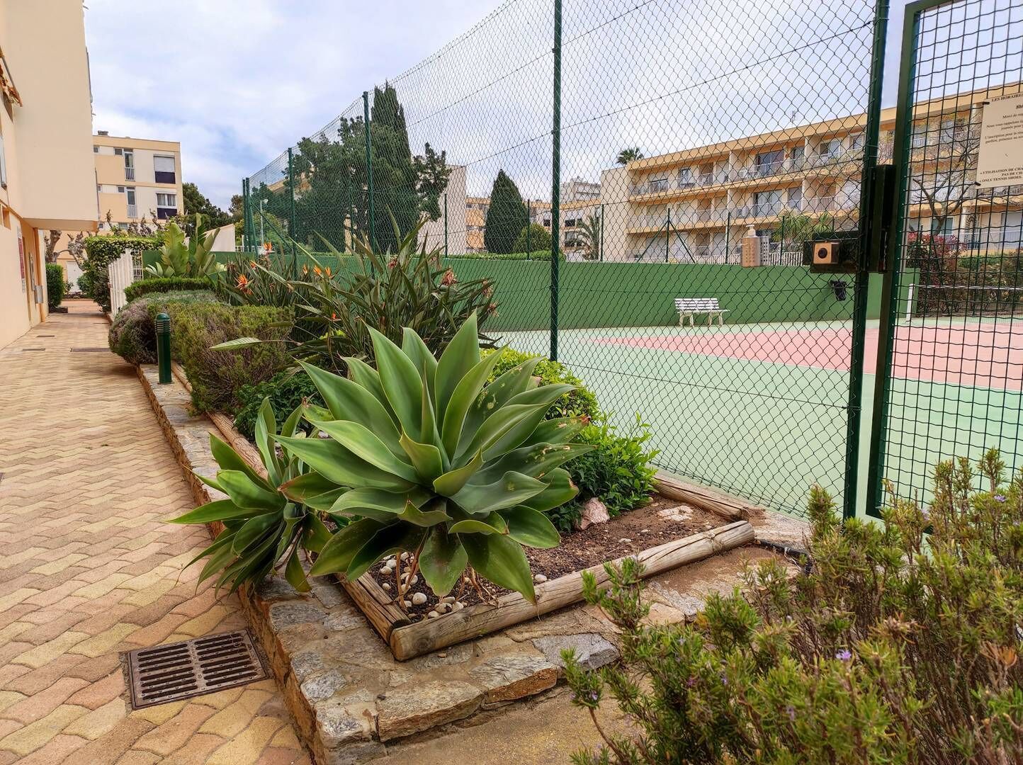 A tennis court surrounded by a green fence, with a bench and a trash bin nearby.