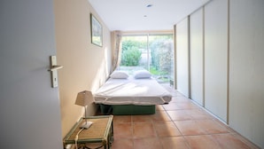 A bedroom with a bed, a bedside table with a lamp, and a view of a garden through a sliding door. - Maison, climatisée plage St Clair et commerces (LE LAVANDOU)