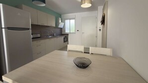 Une cuisine moderne avec un réfrigérateur en acier inoxydable, une table à manger surmontée d’un panier en fil de fer noir et une fenêtre offrant une vue agréable.