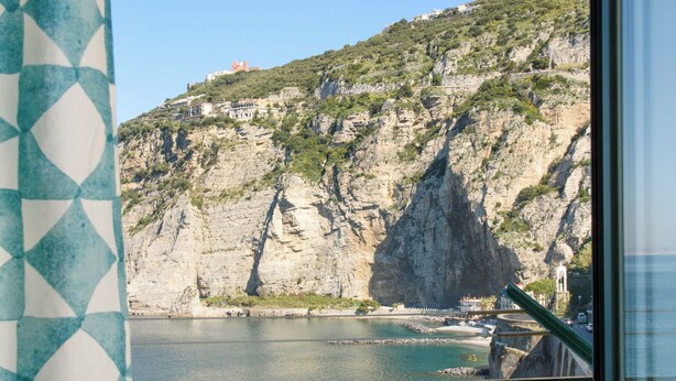 Une vue sur la côte, prise depuis une fenêtre ornée d’un rideau à motifs géométriques, avec une bouteille et une rambarde en métal.