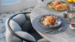 Une table ronde recouverte d’une nappe à motifs bleus et blancs, deux assiettes avec des pâtisseries, un verre d’eau et un vase rempli de fleurs jaunes.
