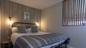 A bedroom with a bed, wooden panel wall, two wall-mounted lights, a window with vertical blinds, and a small stool. - The Hideout-Boutique Cabin, Estuary View & Parking (Salcombe)
