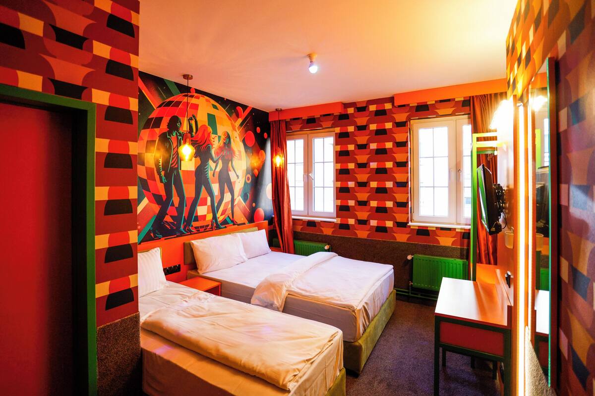 A hotel room with two beds, a small table, and a window with curtains.