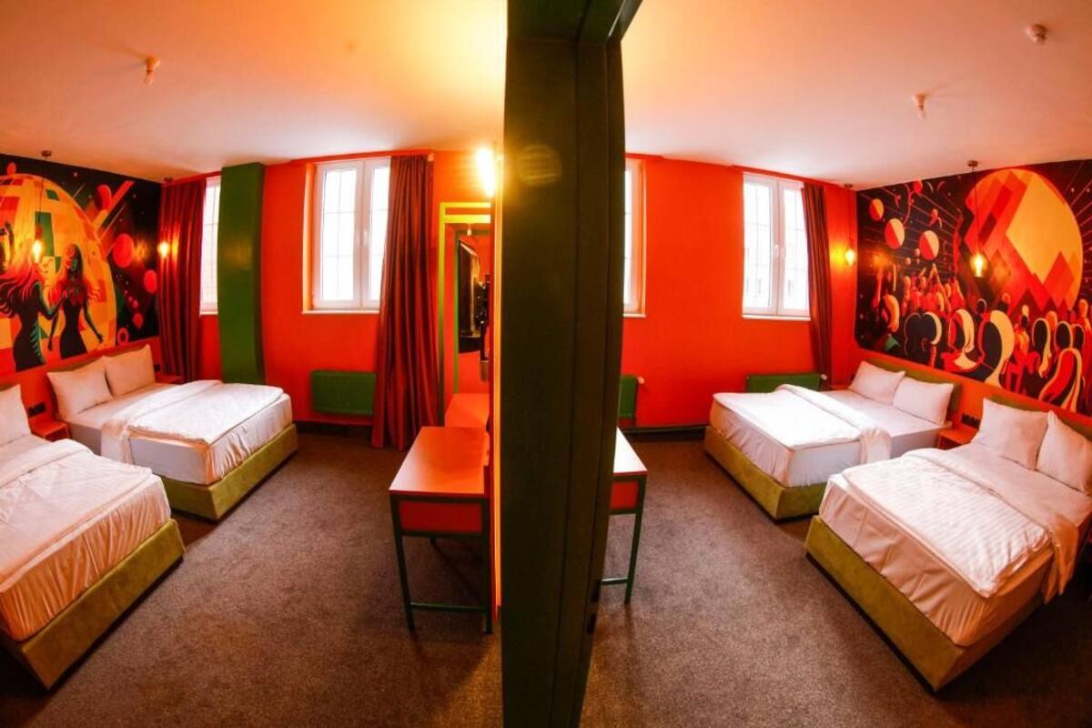 A hotel room with four beds arranged in a U-shape, a dining table, and a vibrant mural on the walls.