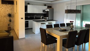 Una cocina moderna con armarios blancos, una isla central y una zona de comedor con mesa de vidrio y sillas negras.